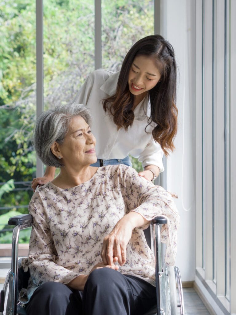 Young Asian caregiver pushing an older woman in a wheelchair in front of corridor with glass wall.